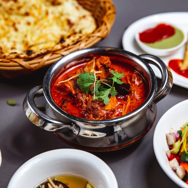 Indian lamb curry in a metal pot with naan bread, chutneys, and salad on a dark table.