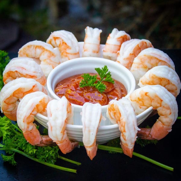 Shrimp cocktail with a white bowl of red sauce and parsley on a dark surface.